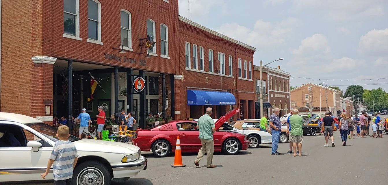 Waterloo IL Optimist Club Car Show on a street with vintage and modern cars, people walking and gathering, brick buildings, a blue awning, and traffic cones.