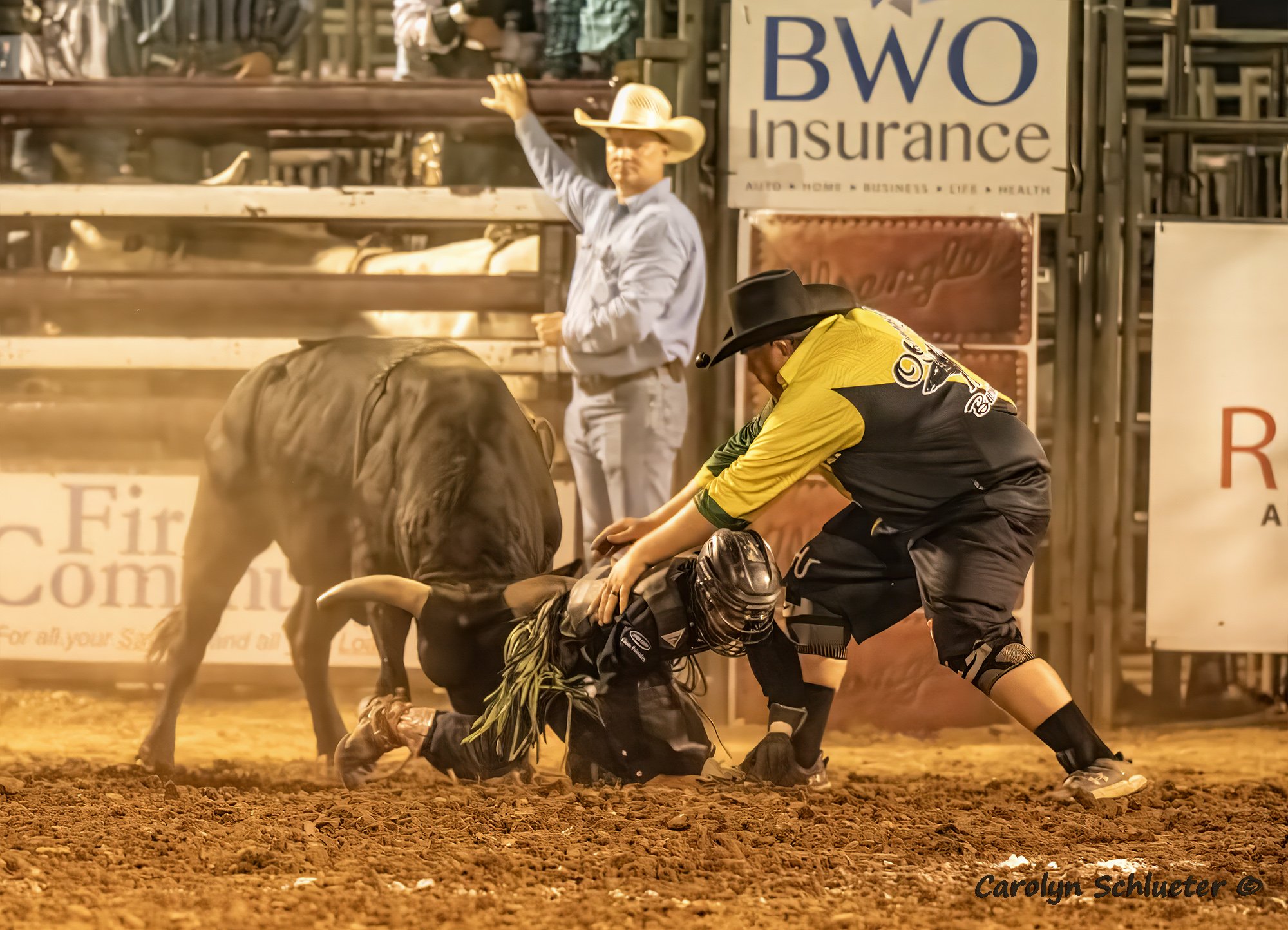 A rodeo scene where a cowboy is assisting a fallen competitor while a bull stands nearby. An announcer in a cowboy hat is in the background, waving to the crowd.
