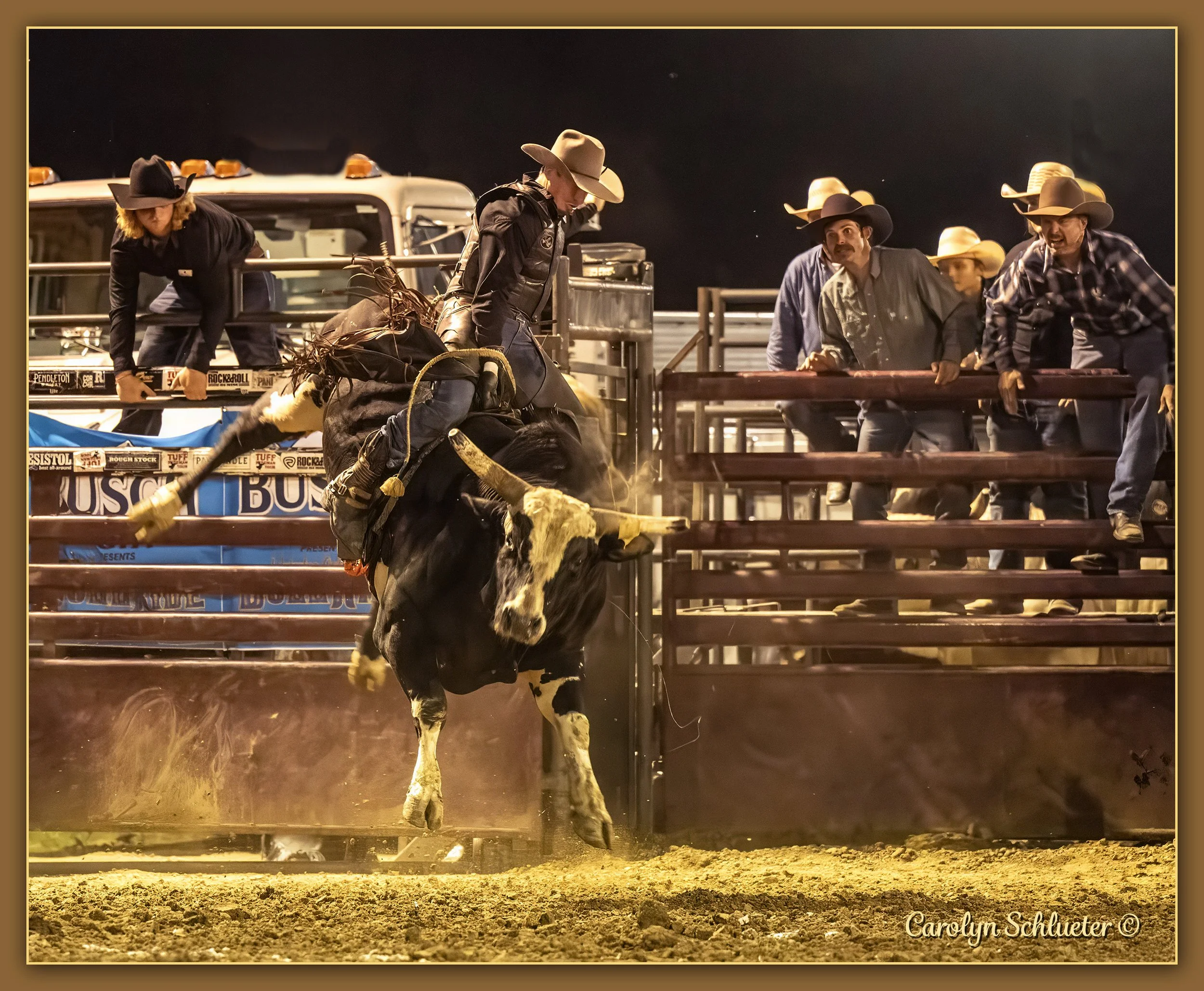 A rodeo scene at the Blue Army Bull Bash with a cowboy riding a bucking bull, surrounded by rodeo officials with cowboy hats watching from the fence.