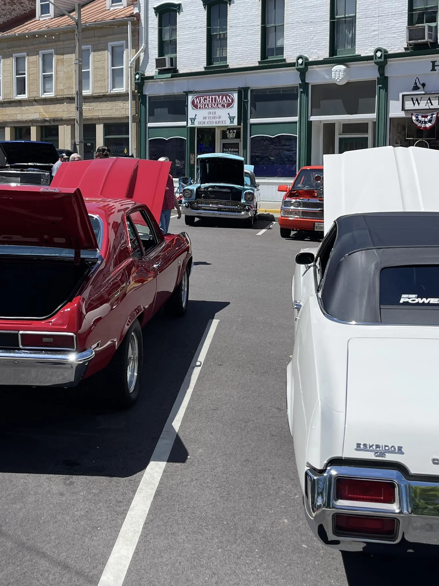 A row of vintage cars parked in front of a pharmacy, with some cars having hoods open at the Optimist Club Car Show The background shows a building with a pharmacy sign, a storefront with large windows, and a few people walking around.