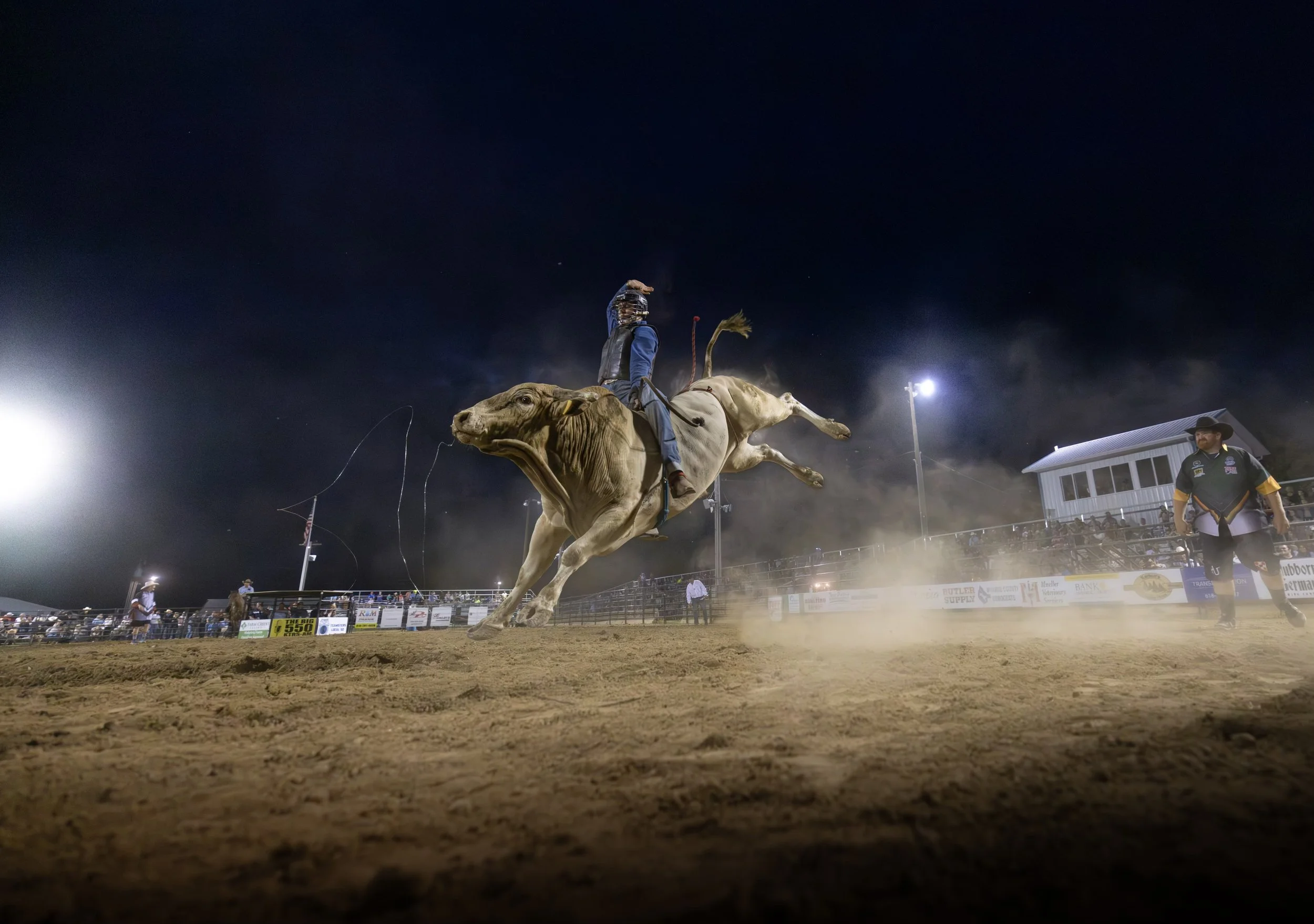Rodeo cowboy riding a bucking bull at night, dust rising on the dirt arena, spectators watching from the stands at the Blue Army Bull Bash.