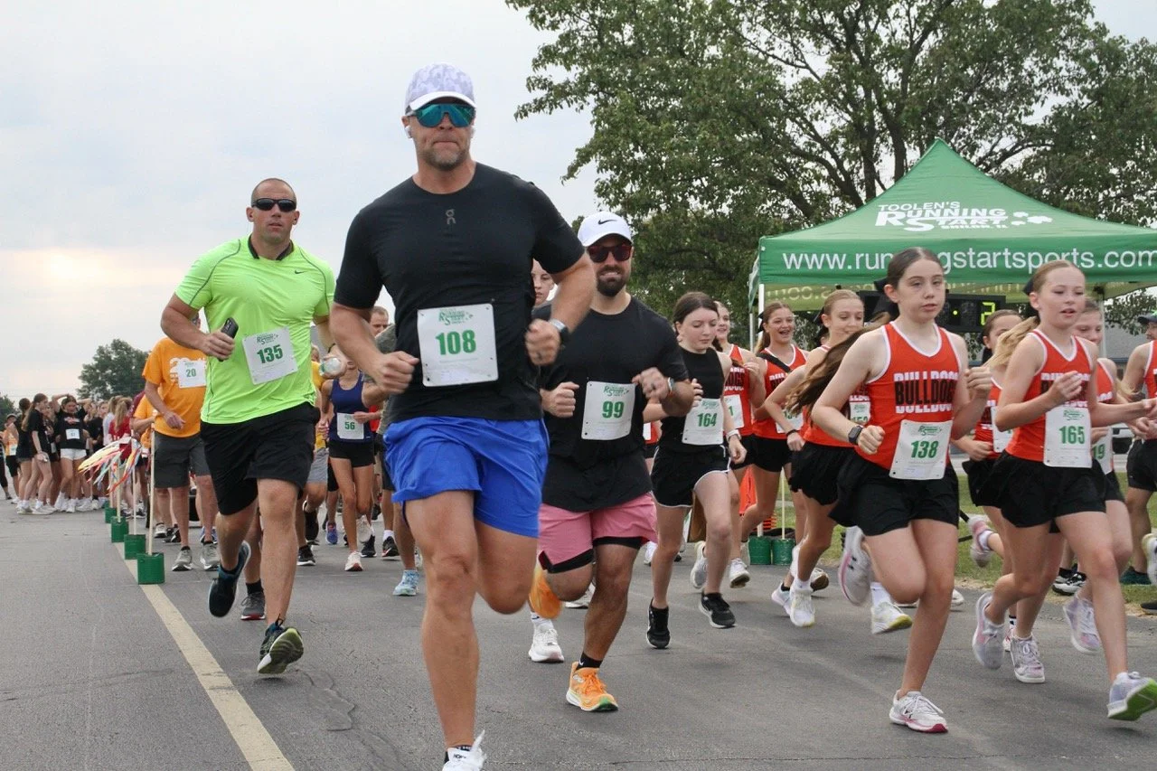 Participants in the Optimist Club 5K and Fun Run, including adults and young girls, running on a street with a green tent in the background. The runners wear various athletic outfits and race bibs.