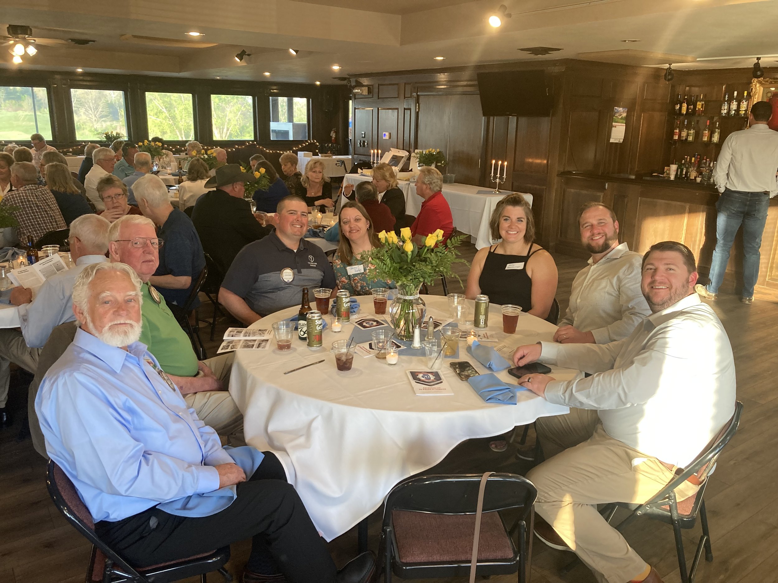 A group of people sitting around a table at a social event or celebration in a well-lit banquet hall, with drinks and a flower centerpiece on the table, and other guests in the background.