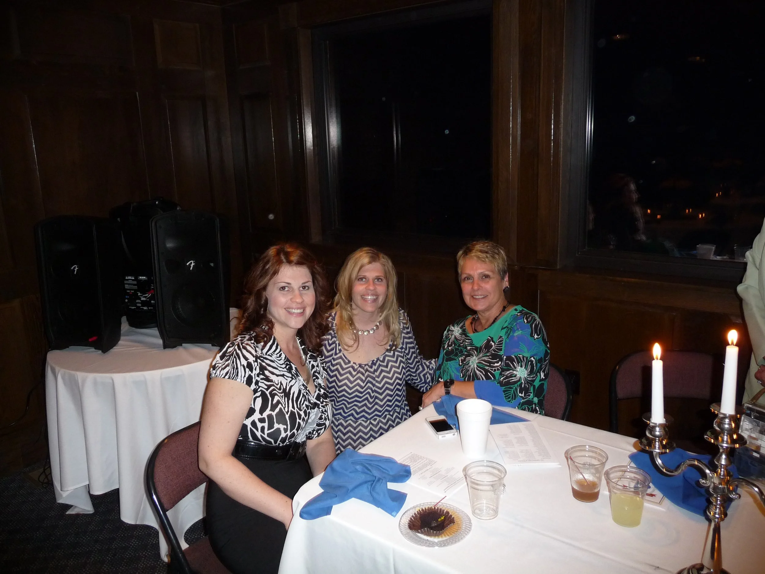 Three women sitting at a round dinner table, smiling at the camera. The table has a white tablecloth, blue napkins, candles, drinks, and papers. There are speakers and a window in the background.