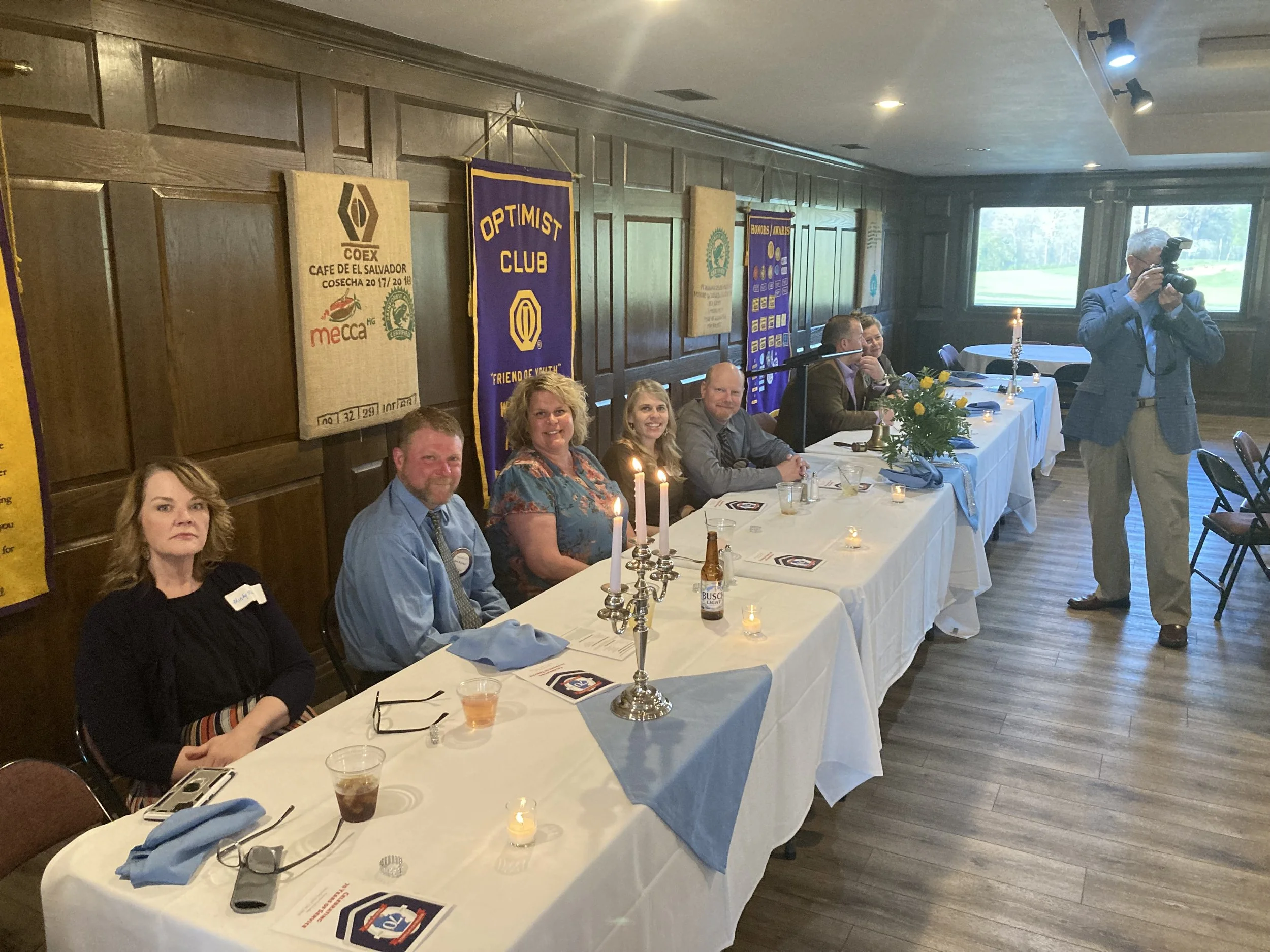 A group of people sitting at a banquet table during a formal event, with banners on the wall behind them and a man taking a photograph.