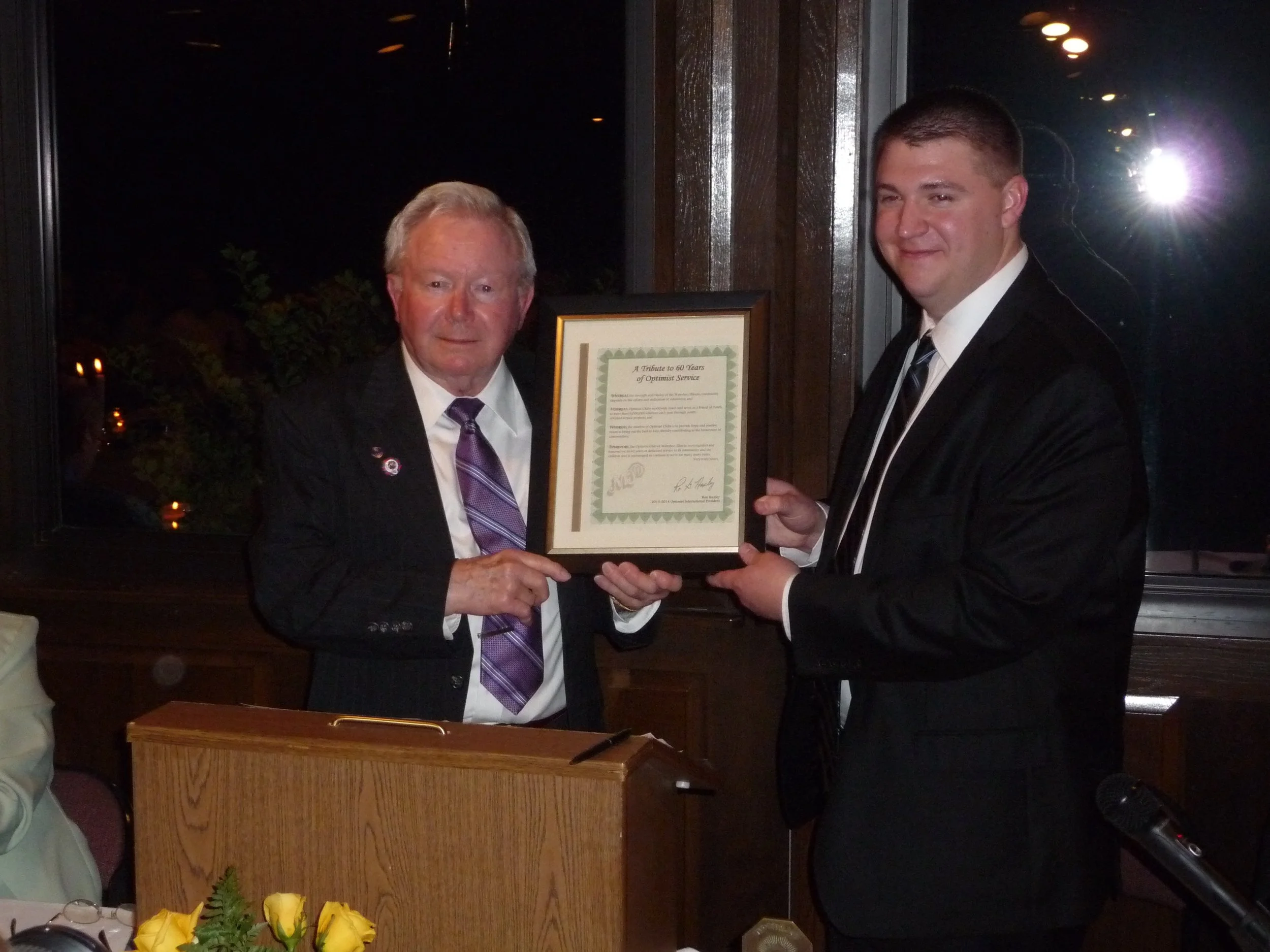 An older man in a black suit with a purple tie and a younger man in a black suit with a striped tie stand indoors at night, holding a framed certificate together at the Waterloo Optimist Club anniversary celebration. 
