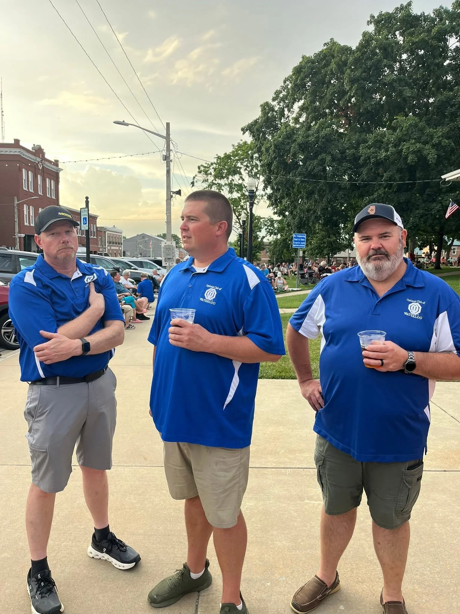 Waterloo Optimist Club members on a concrete sidewalk, holding drinks, with people sitting on benches and a park and brick building in the background.