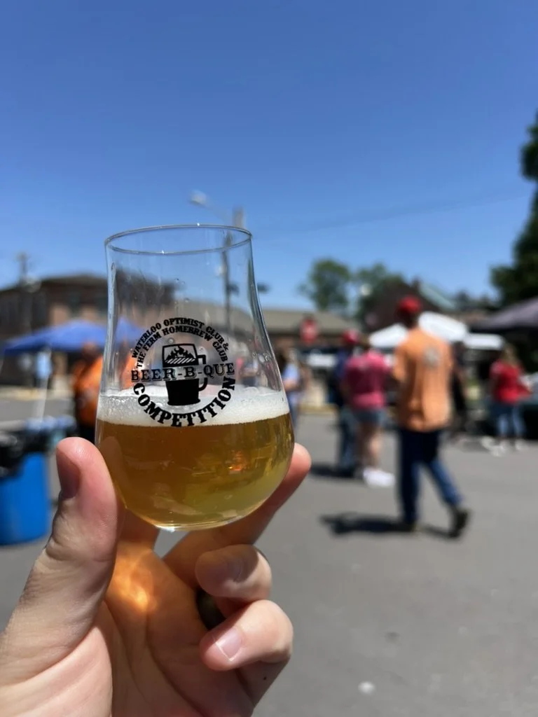Person holding a Beer-B-Que beer glass with a logo at an outdoor Waterloo Optimist Club event with people walking around, tents, and blue sky in the background.