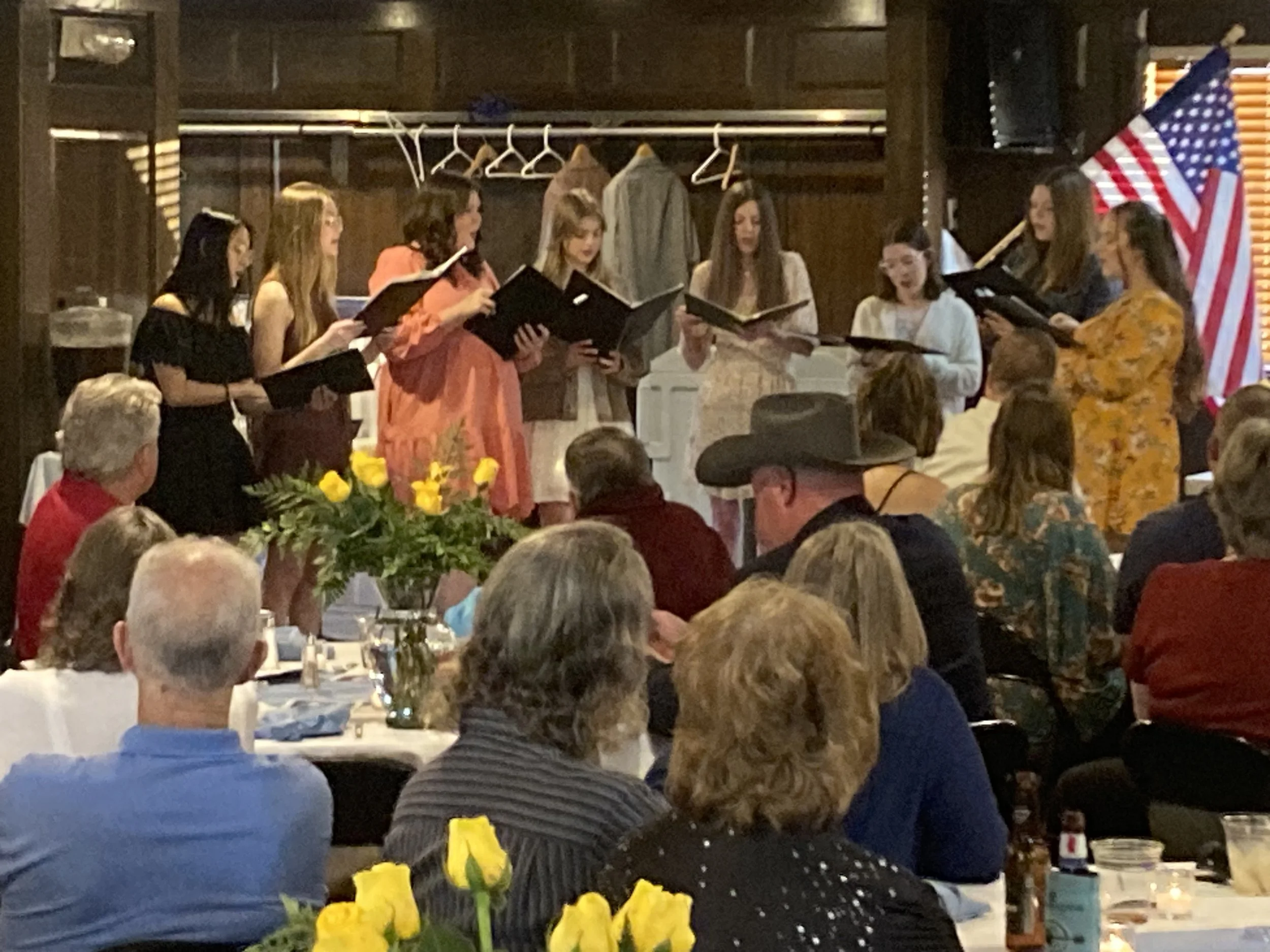 A group of women singing in a choir performance indoors, with an American flag in the background and an audience seated at tables decorated with yellow flowers.