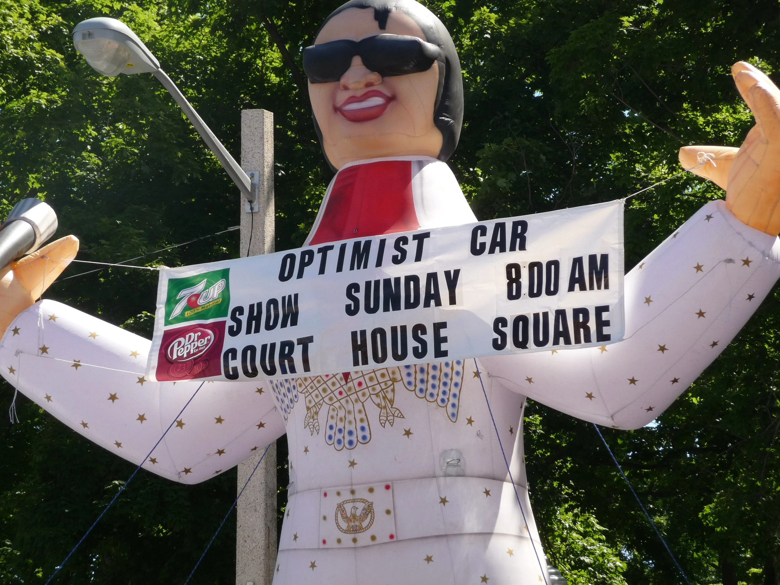 Inflatable figure of a woman with black hair, sunglasses, wearing a white dress with gold stars and eagle emblem, holding a sign with Sprite, 7UP, and Dr Pepper logos advertising the Waterloo Optimist Club car show.