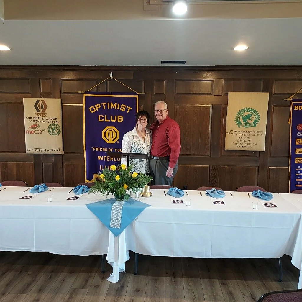 Two people standing behind a long banquet table, smiling at the camera, with banners hanging on the wood-paneled wall behind them, including an Optimist Club banner, in a room decorated for a formal event.
