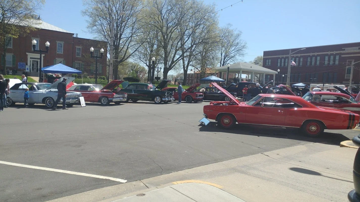 A row of classic cars on display at the Waterloo IL Optimist Club Car Show, with open hoods and people observing them on a sunny day in a town square.