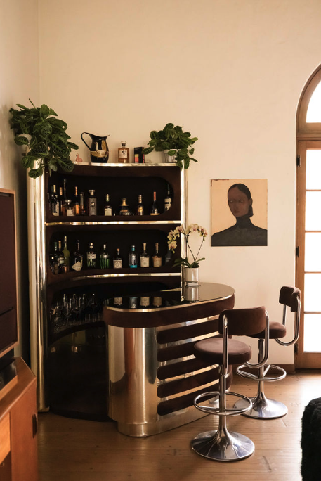 An indoor home bar with a curved wooden counter, two bar stools, a dark wooden shelf filled with liquor bottles, potted plants, and a framed portrait on the wall near an arched window.