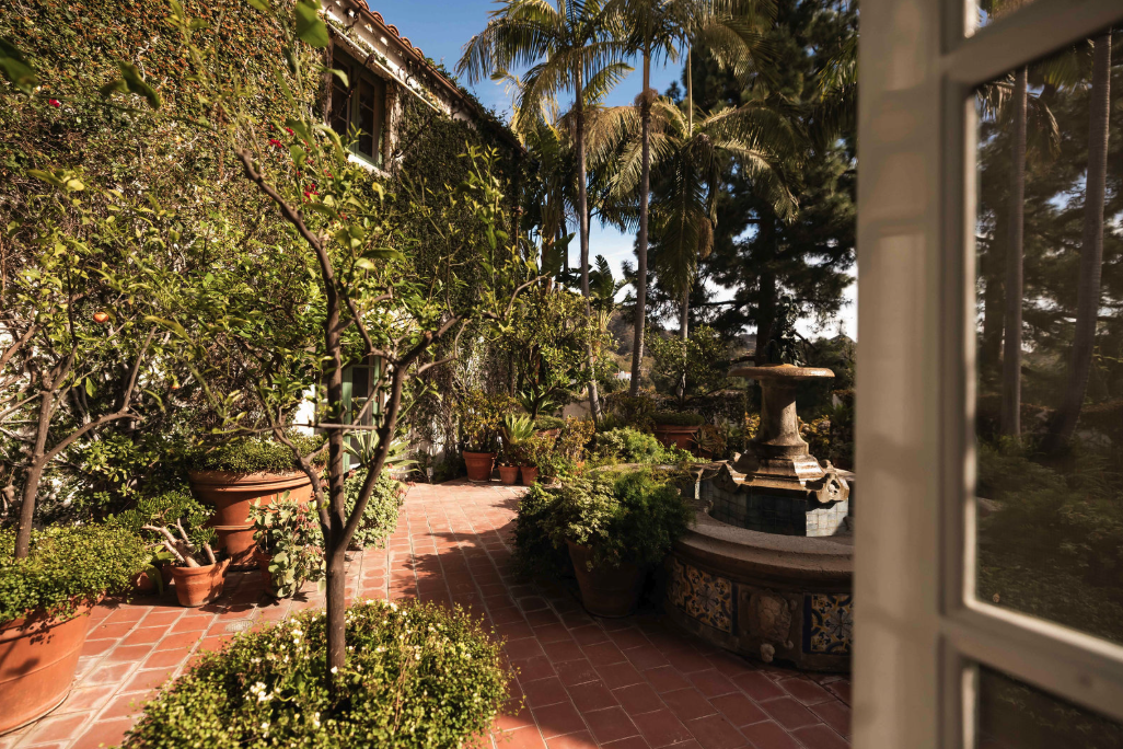 A lush outdoor patio with potted plants, a fountain, brick flooring, and tall palm trees, viewed from an open window.