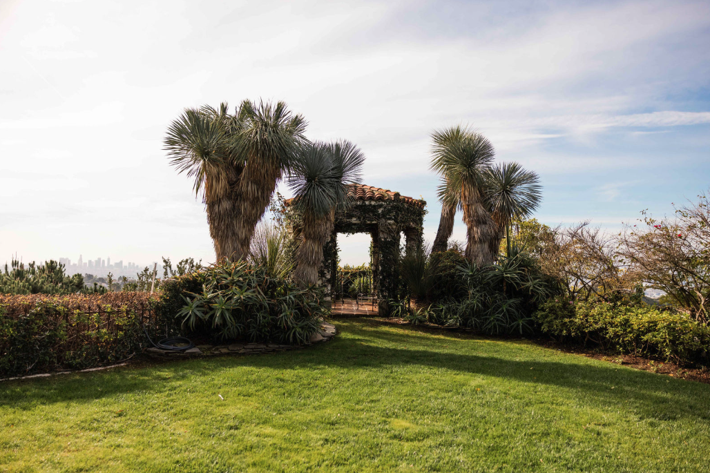 A garden area with lush green grass, tall palm trees, bushes, and a small wooden gazebo with a tiled roof.
