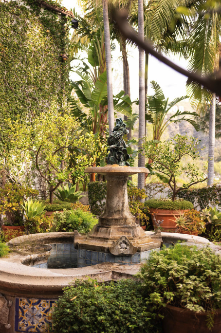 A decorative water fountain featuring a lion sculpture in a lush garden with palm trees and potted plants.