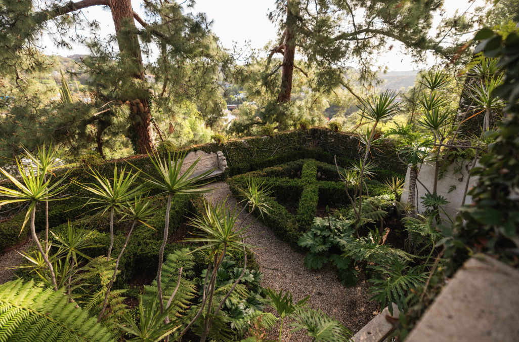 A lush garden with a gravel pathway winding through manicured hedges, featuring a cross-shaped hedge and surrounded by various leafy plants and tall trees.