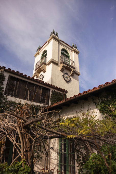 A white clock tower with a red tiled roof and green windows, partially obscured by a building with a similar roof, under a partly cloudy sky, with dried branches and green foliage in the foreground.