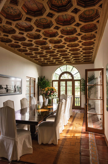 Elegant dining room with a long wooden table surrounded by white upholstered chairs, large floral centerpiece, arched window, and open double doors leading to a balcony, with a decorative ceiling and sunlight streaming in.