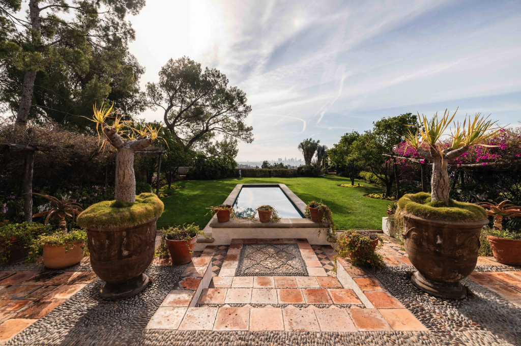 View of a backyard garden with potted plants, a rectangular pool, lush green lawn, and trees under a partly cloudy sky.
