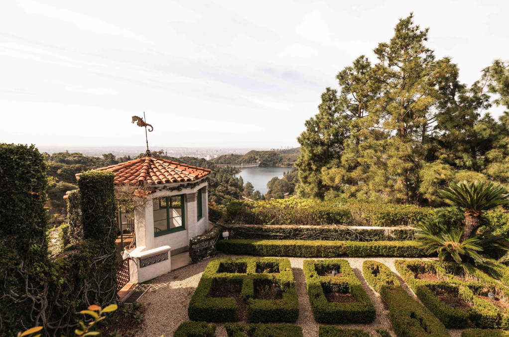 A scenic view of a garden with topiary shrubs, a small white building with a red tile roof topped with a seahorse weather vane, and a large body of water in the background surrounded by trees and hills.