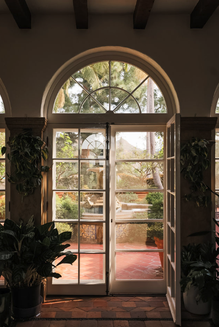 View through glass-paneled double doors with an arched window above, leading to an outdoor patio with trees and plants.