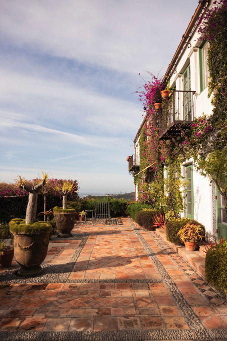 Terrace with potted plants, flowering vines, and a white building with green shutters and balcony, overlooking a scenic landscape.