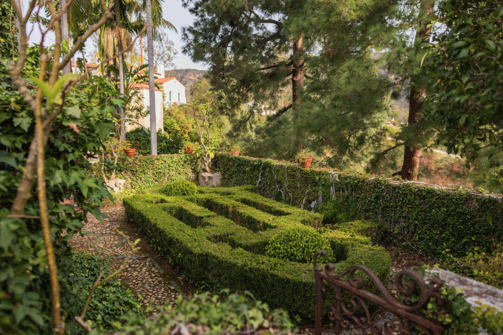 A manicured garden with trimmed hedges, stone pathway, and potted plants, surrounded by tall trees and lush greenery.