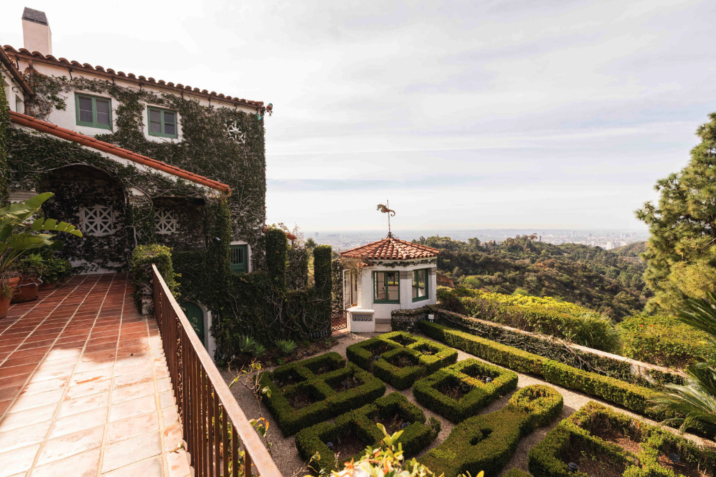 Terrace with potted plants overlooking a garden with shaped hedges and a small structure with a weather vane, surrounded by hilly landscape.