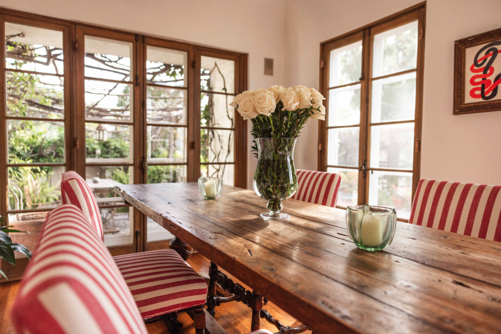 A dining room with a rustic wooden table, pink and white striped upholstered chairs, glass candle holders, a glass vase with white roses, large windows with wooden frames, and a wall art piece.