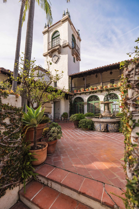 A courtyard with potted plants, a stone fountain, and a white Spanish-style building with a tower and balcony, set amidst greenery and palm trees under a blue sky.