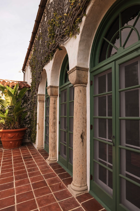 Exterior view of a building with three arched windows with green frames, beige columns, a tiled patio, and green plants in a pot.