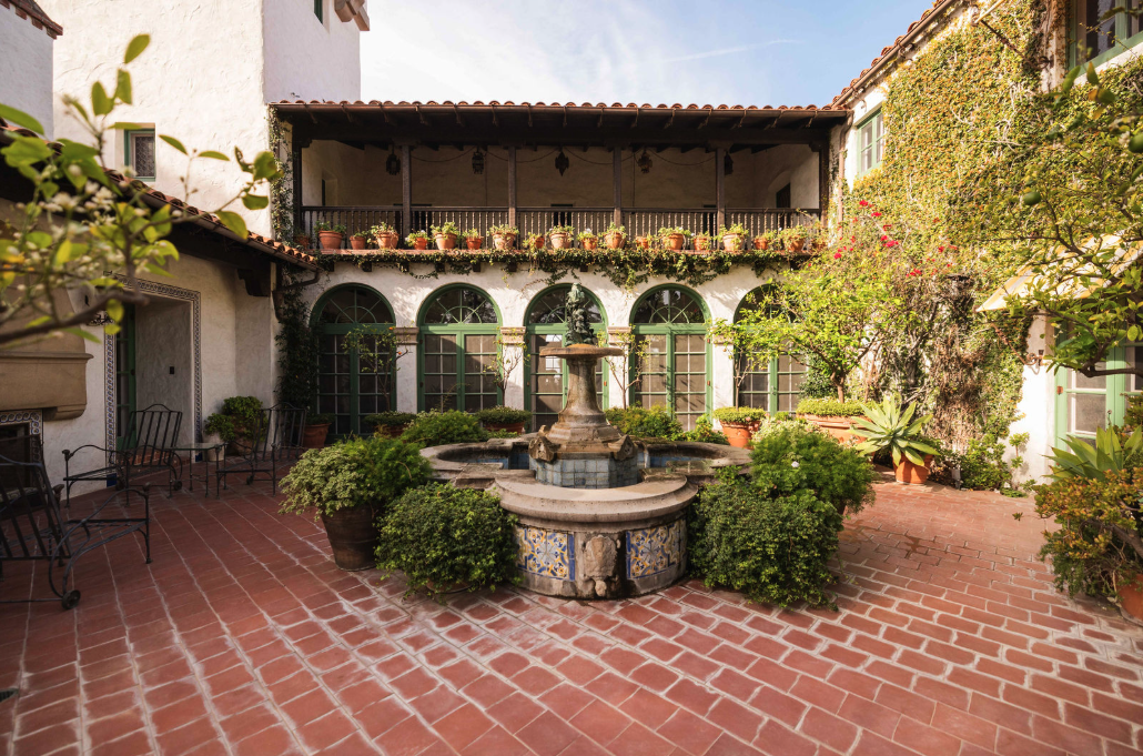 Courtyard with a fountain, potted plants, green foliage, and an arched balcony in a Spanish-style building.