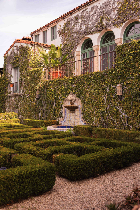 A garden with neatly trimmed hedges, a stone fountain with a lion sculpture, and a building facade covered in green vines and plants.