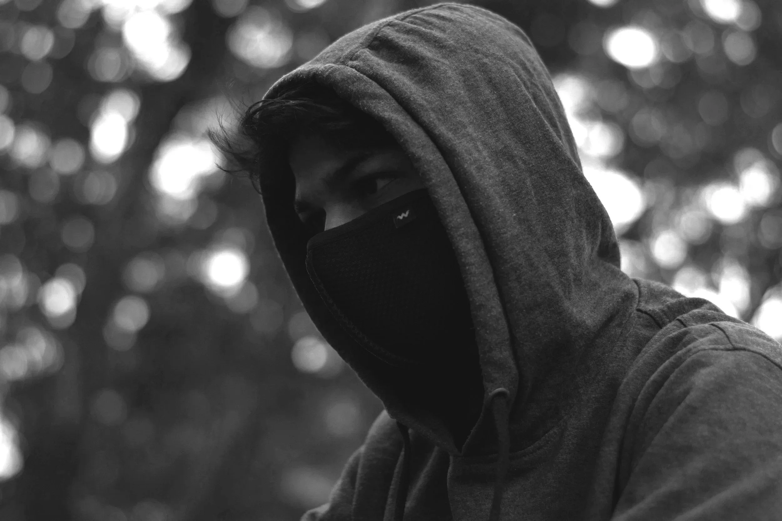 A person wearing a hoodie and face mask outdoors, with blurred trees and bokeh lights in the background.