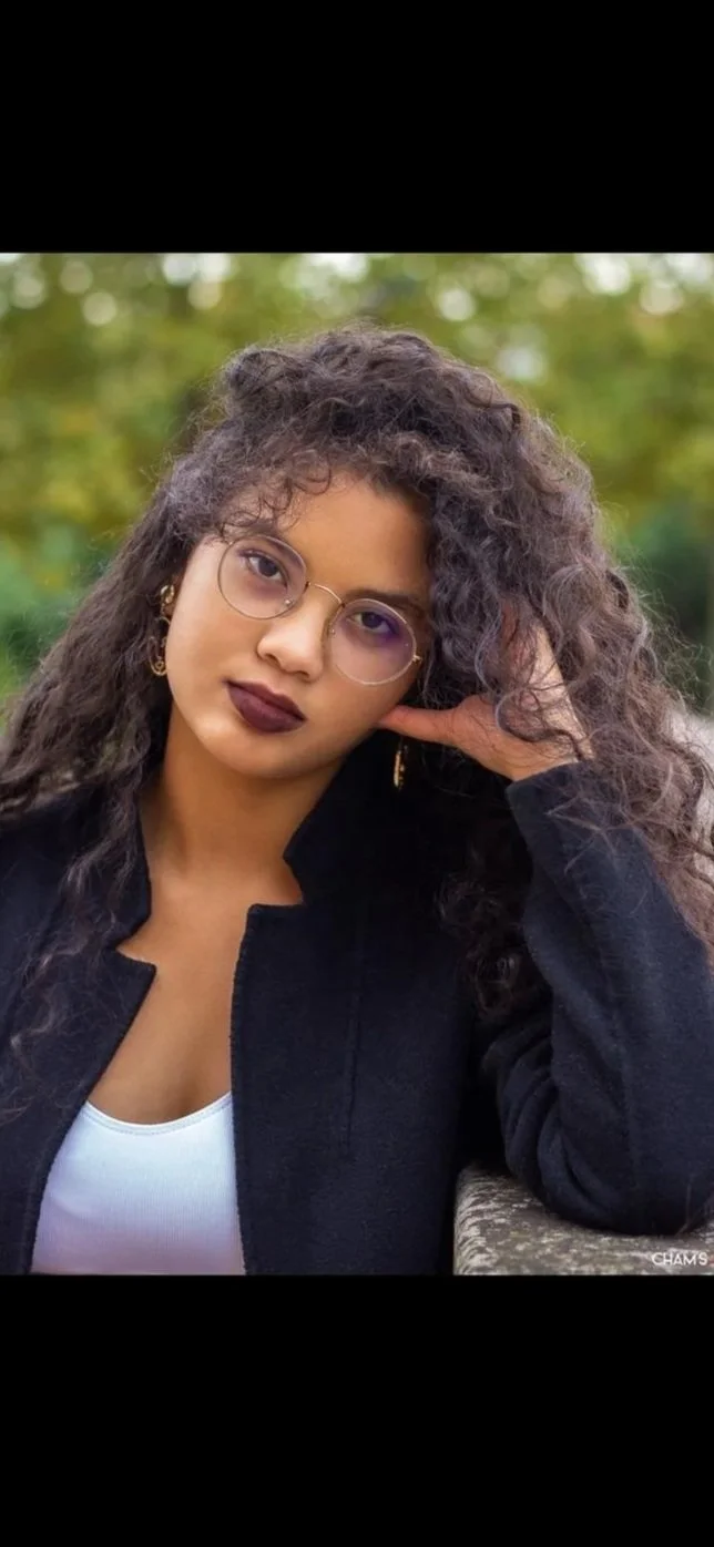 A young woman with curly hair and glasses, wearing dark lipstick, a white top, and a black jacket, sitting outdoors with a blurred green background.