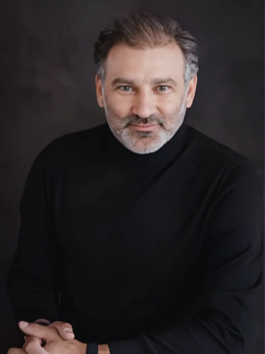 A middle-aged man with gray hair and a beard, wearing a black turtleneck, posing against a dark background.
