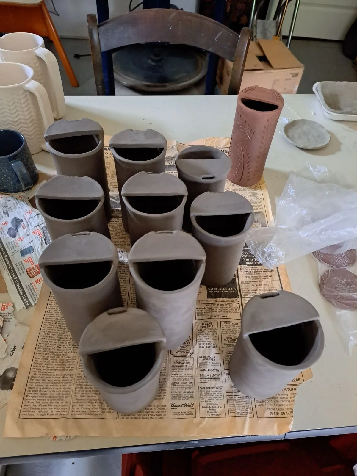 Multiple unglazed clay planters and a pink ceramic vase on a table covered with newspaper, with pottery supplies and a box in the background.