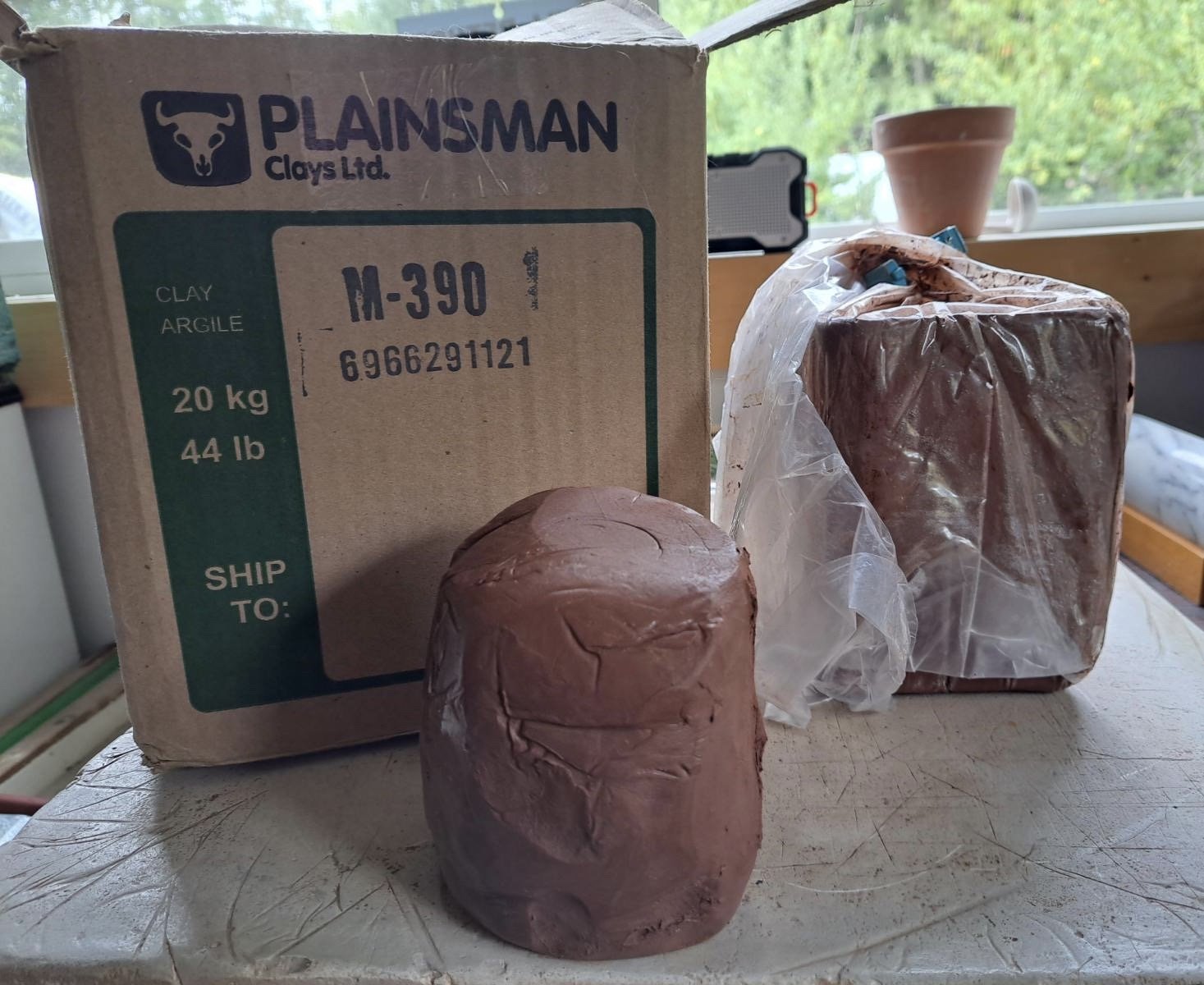 A cardboard box labeled 'Plainsman Clays Ltd.' with clay blocks and a piece of clay on a table in front of a window.