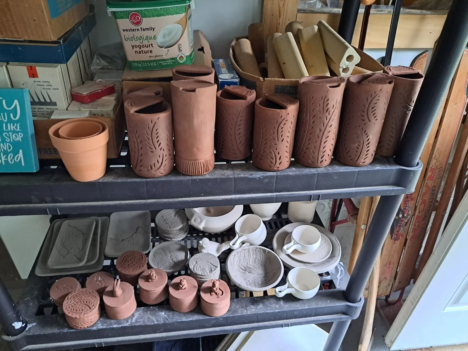 Shelves filled with various unfinished ceramic pottery items, including tall vases, cups, plates, and small containers, some with carved patterns and some plain.