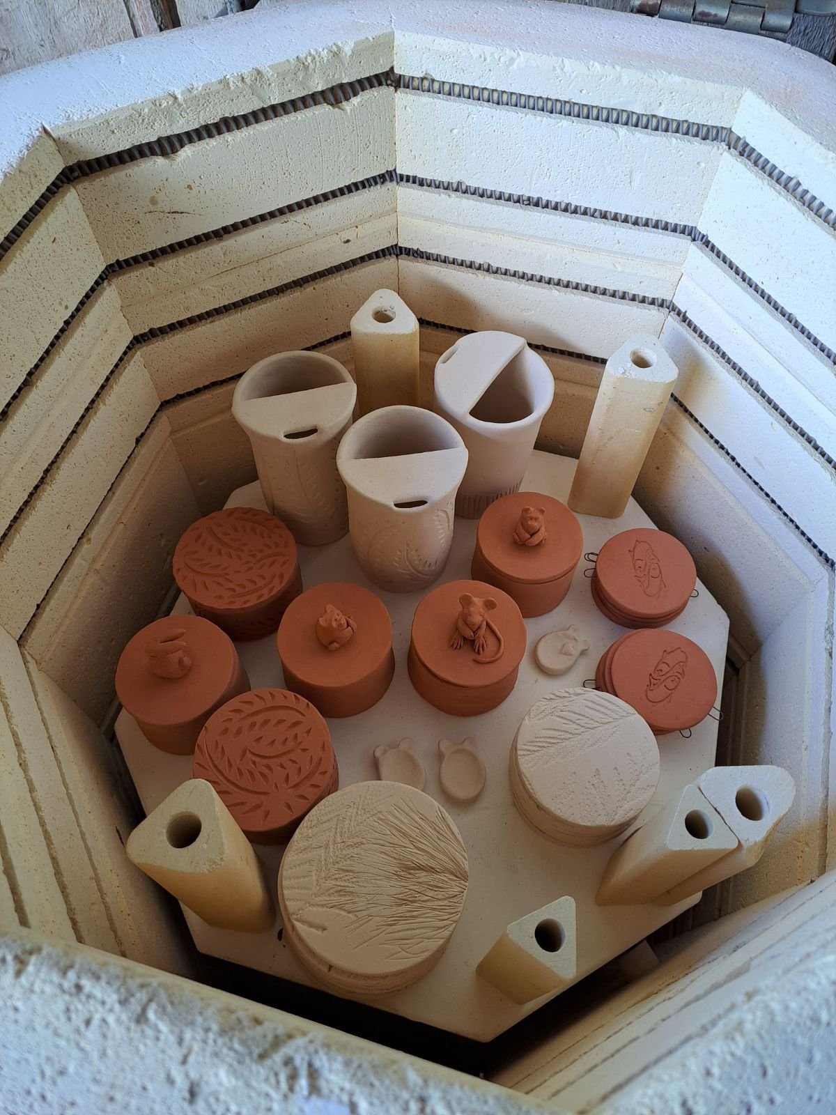 A collection of ceramic and clay pottery items, including cups, lids, and abstract designs, arranged inside a brick and stone-lined kiln or oven.