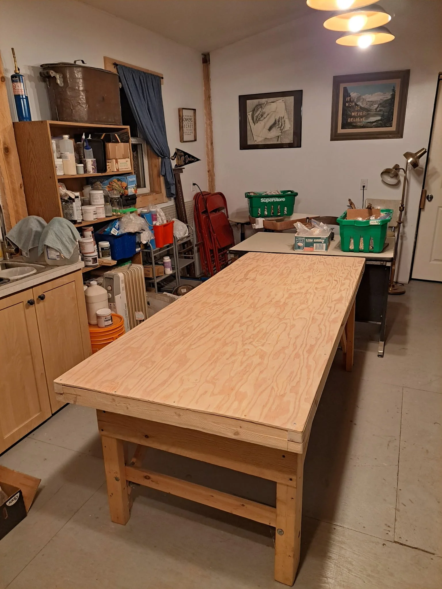 A large, unfinished wooden workbench in a basement or workshop with shelves containing paint, tools, and supplies, red chairs stacked against the wall, framed pictures, and a small table with green plastic baskets.