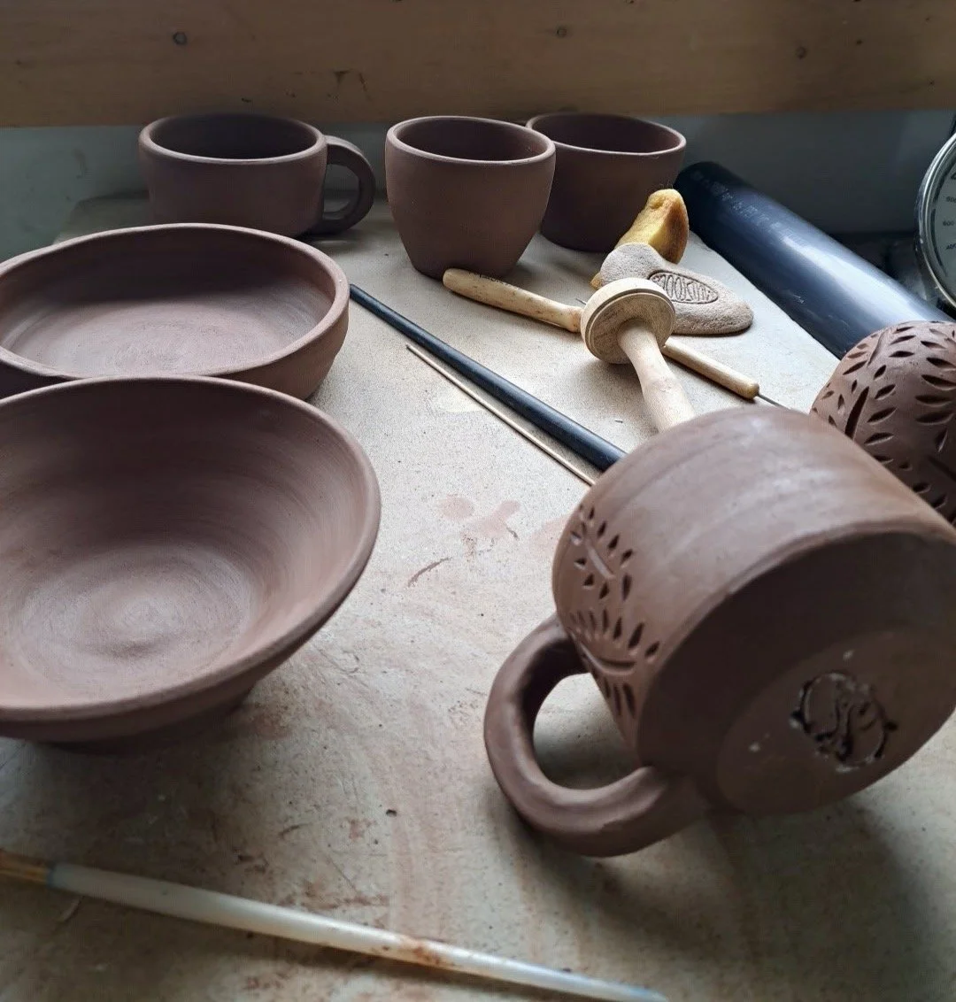 Unfinished pottery pieces, including bowls and mugs, on a worktable with sculpting tools and a speedball, indicating a pottery or ceramics workspace.