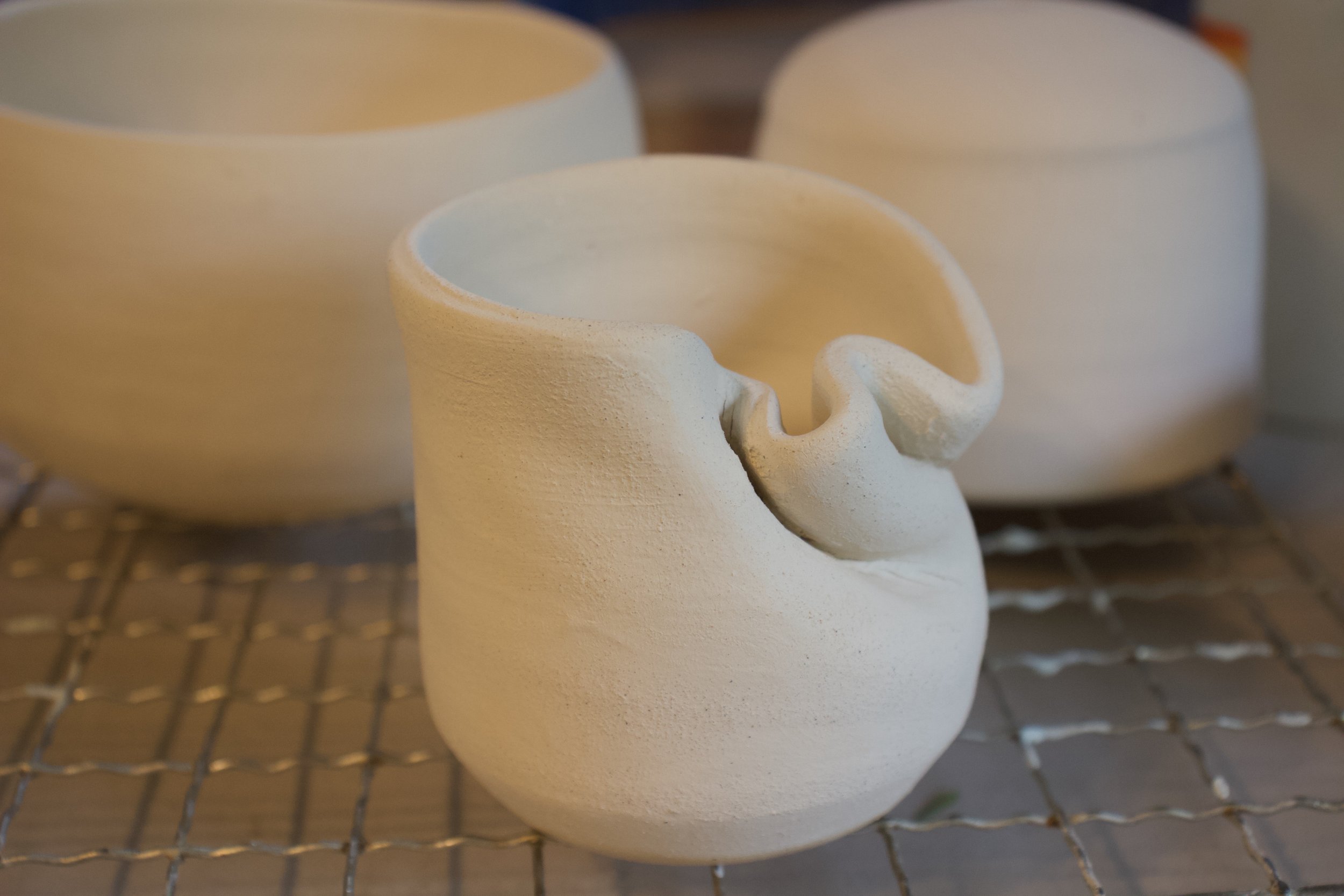 White ceramic cups with a textured surface, one shaped with a handle resembling a curled finger, on a wire rack.
