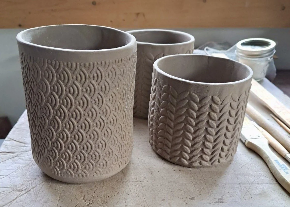 Three unfinished clay pots with textured patterns on a worktable, with pottery tools and a jar in the background.