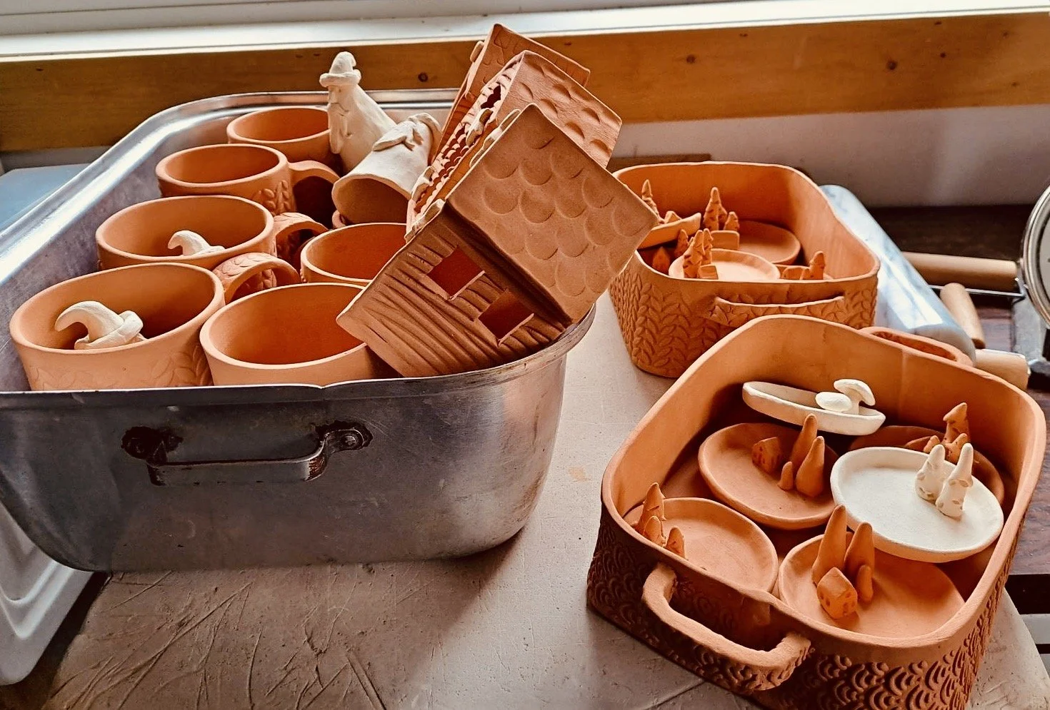 Unfinished pottery pieces, including cups, bowls, and figurines, arranged in trays and a metal container on a table in a ceramics studio.