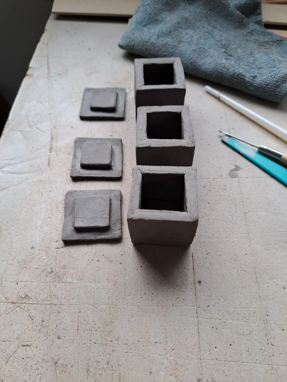 Several unfinished gray concrete square pots with separate square lids on a work surface, with a towel and pens nearby.
