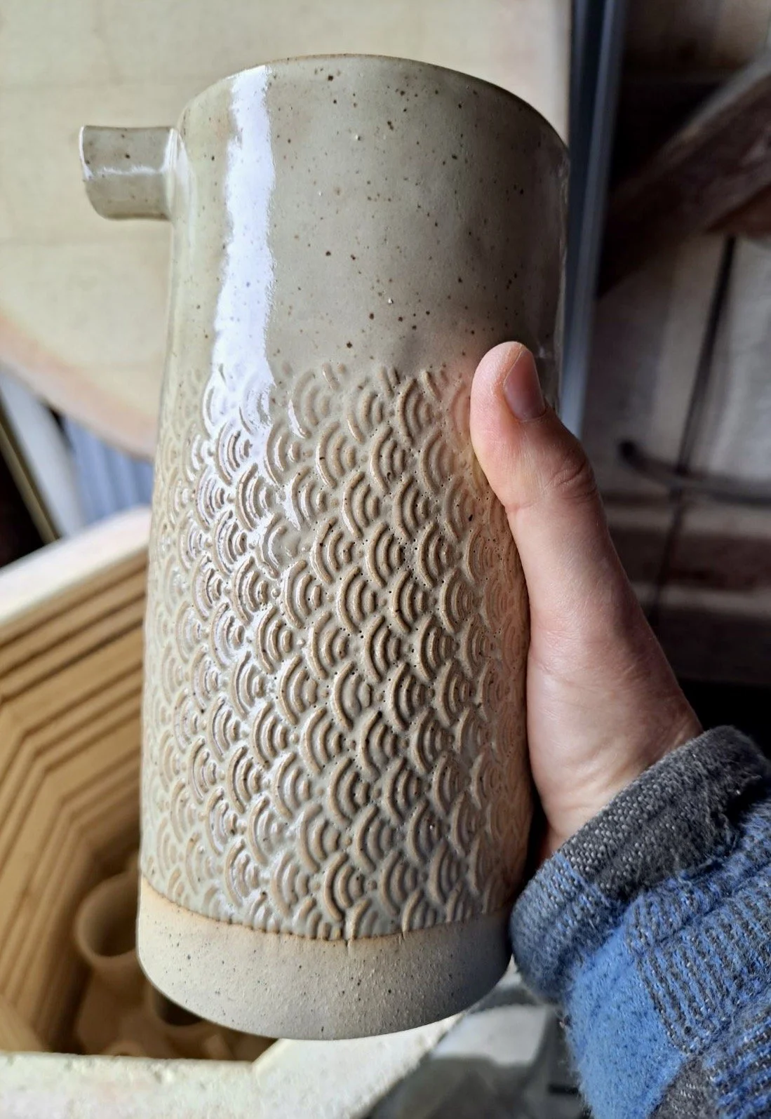 Hand holding a beige ceramic pitcher with a textured pattern and a small spout, against a background of wooden surfaces and a crate of other ceramics.
