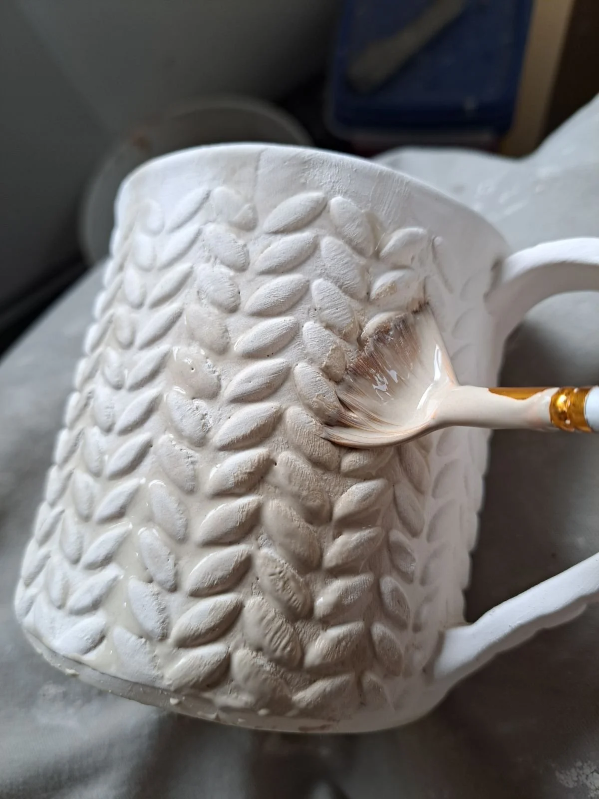A ceramic mug with a textured leaf pattern, being painted with a small brush dipped in white paint.