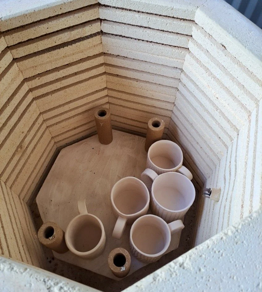 Six ceramic coffee mugs placed inside a kiln, with empty shelves and brown kiln supports.